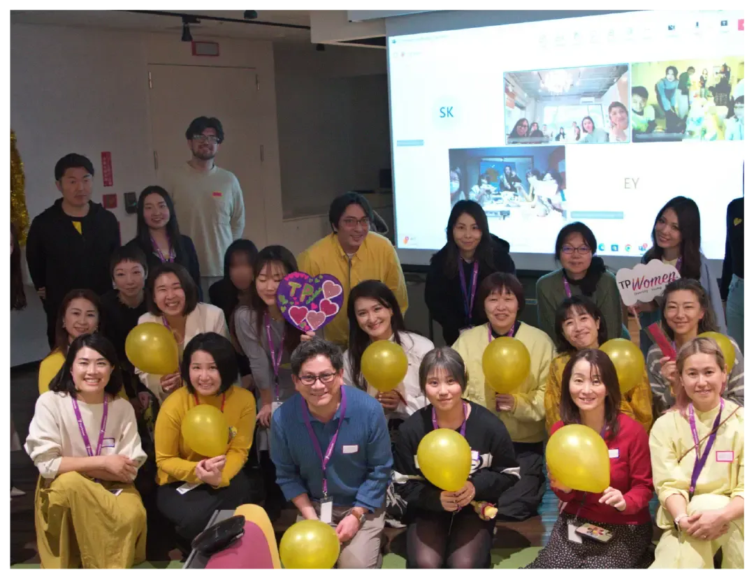 A diverse team celebrates together at a TP Women event with yellow balloons.