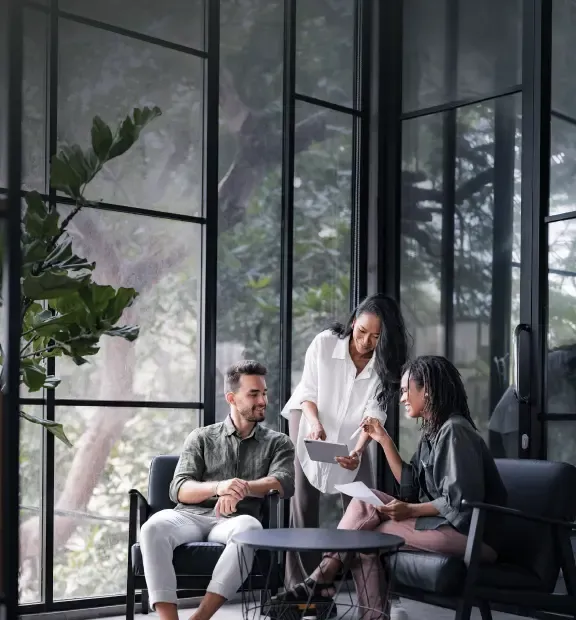 Three colleagues collaborate cheerfully in a bright modern office, using a tablet and discussing ideas.
