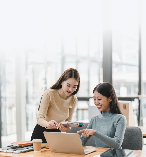 Two professional women collaborate happily at work, discussing ideas on a laptop in a bright modern office.