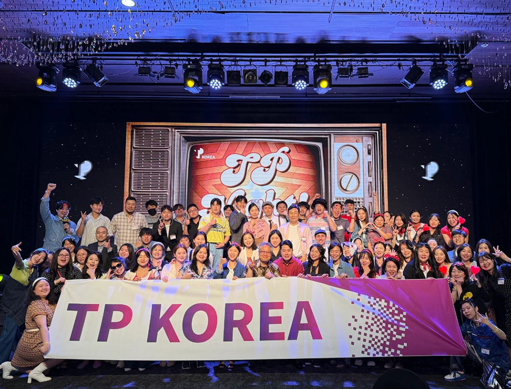 A large group of TP Korea employees pose cheerfully with a banner at a company event.