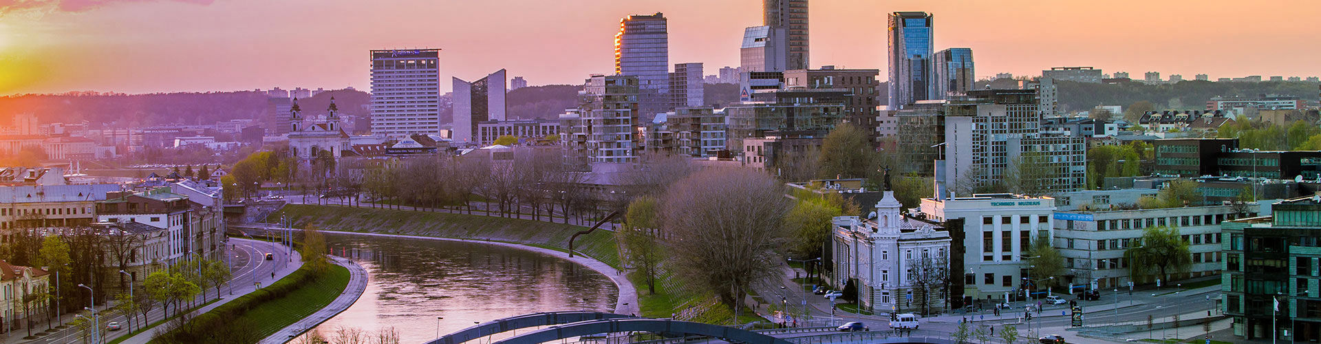 Panoramic view of Vilnius, Lithuania, at sunset, featuring the Neris River, historic buildings, tree-lined streets, and the modern city skyline.