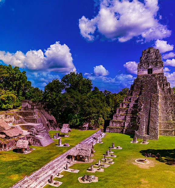 Panoramic view of the ancient Maya ruins at Tikal, Guatemala, surrounded by lush tropical forest, with towering stone temples, plazas, and historic structures under a partly cloudy sky.