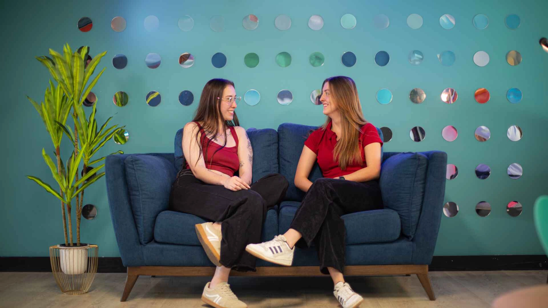 Two women sitting in a blue couch smiling at eachother.