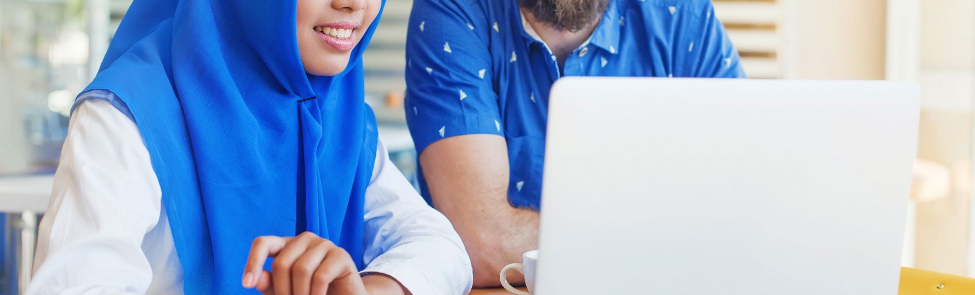 Woman wearing a blue hijab collaborating with a colleague while working on a laptop.