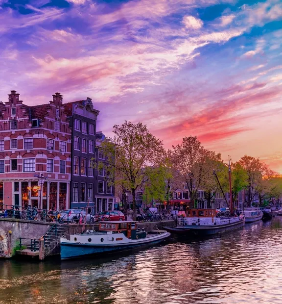 Canal view in Amsterdam, Netherlands, at sunset, featuring historic narrow houses, bicycles, boats, and tree-lined streets.