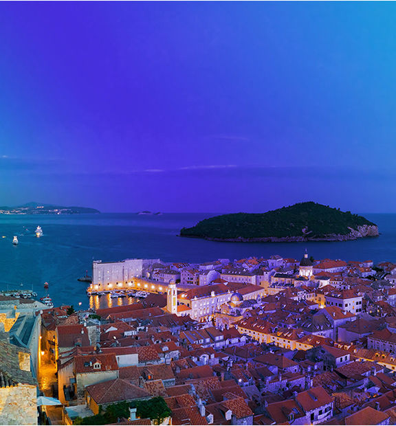 Aerial view of Dubrovnik's historic old town in Croatia at dusk, with the walled city's red-tiled rooftops illuminated, the old harbor below, and the forested Lokrum island in the background under a deep blue-violet sky.