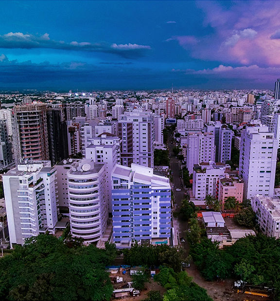 Aerial panoramic view of a dense urban skyline in the Dominican Republic at dusk, showing rows of high-rise apartment and office buildings under a dramatic sky transitioning from deep blue to pink and purple clouds.