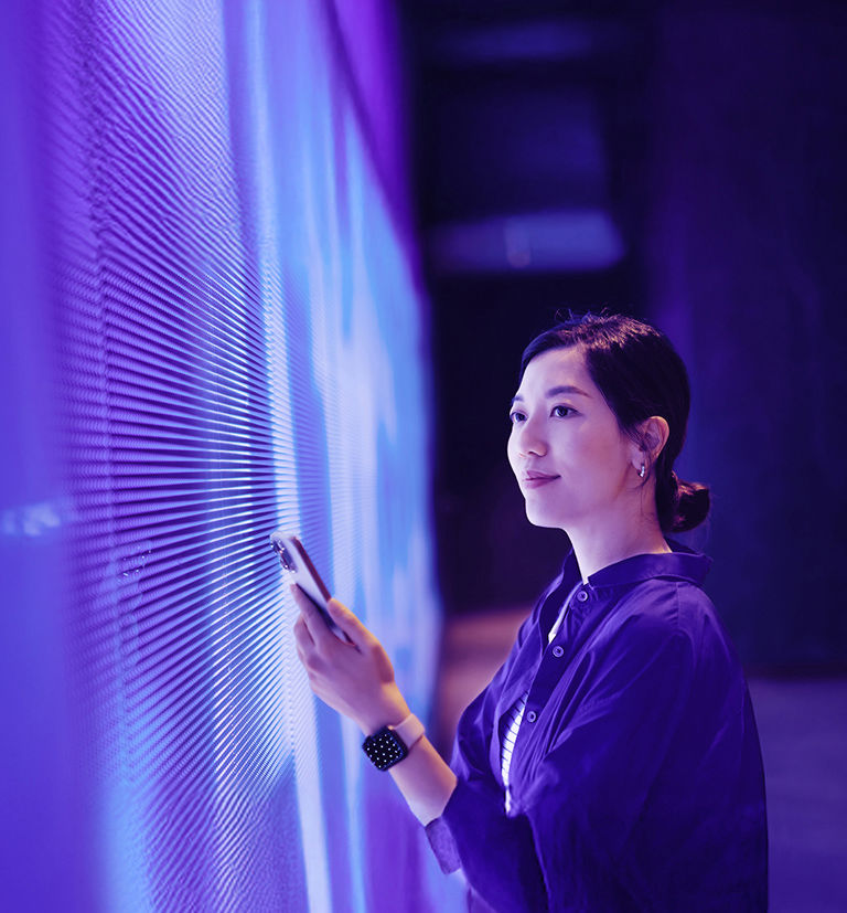 Asian woman holding a smartphone in front of a modern digital display.