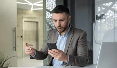 Man victim of financial crime looking at his cell phone in surprise while holding his credit card