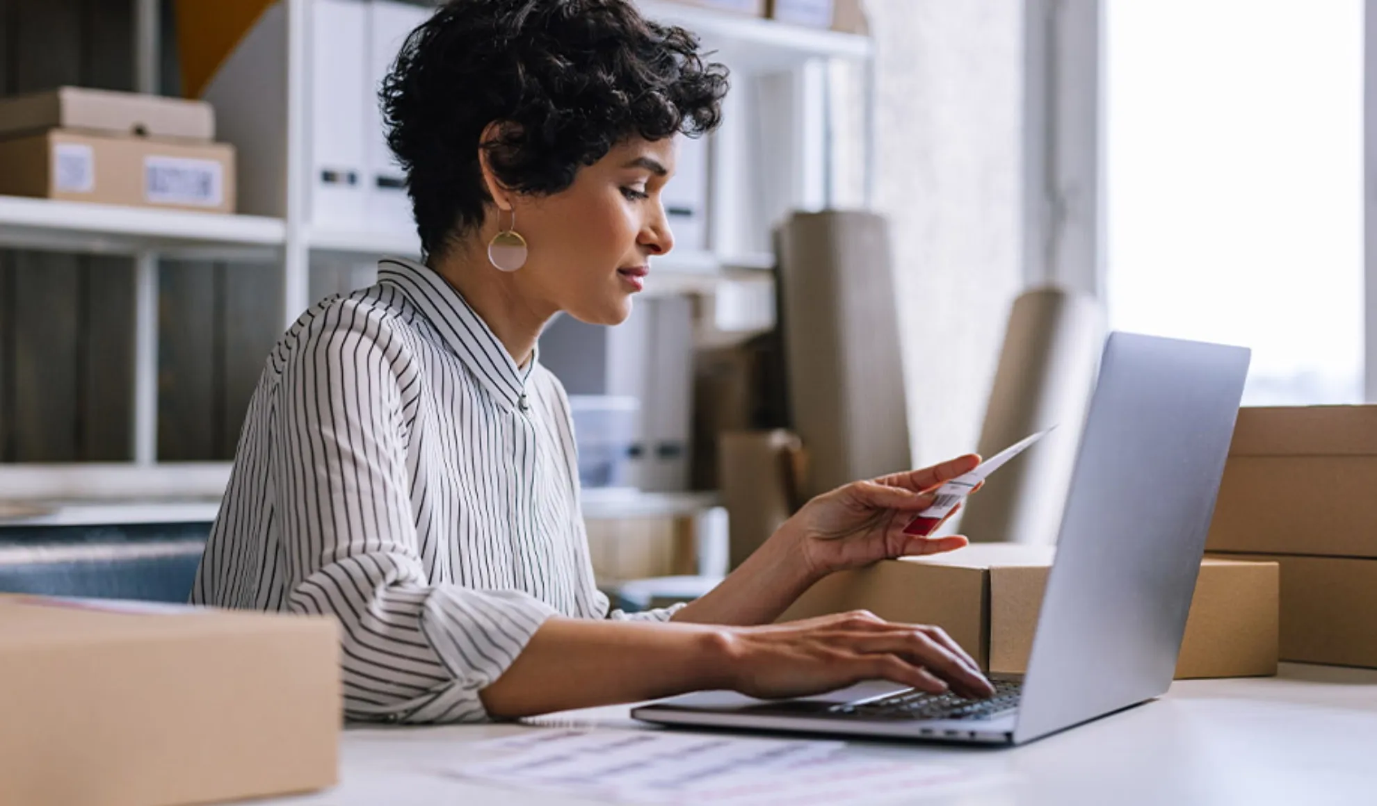 Businesswoman holding a receipt while working on a computer among delivery boxes.