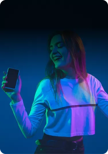 a person holds a smartphone and dances joyfully, illuminated by vibrant pink and blue lights against a dark background.