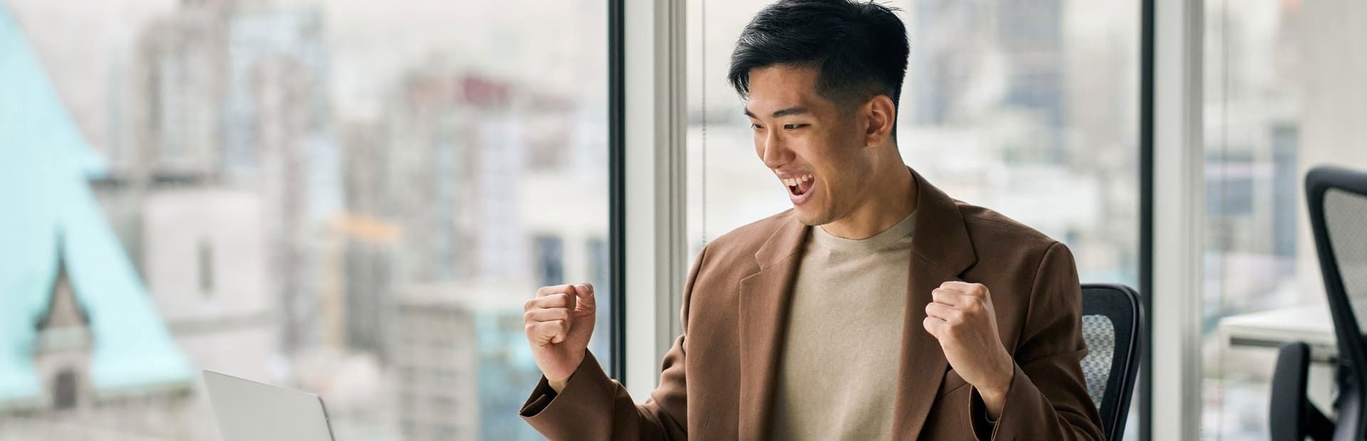 Asian man in a brown suit celebrates in front of his computer after receiving good news, inside a modern glass-walled office.