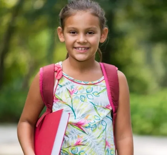 Young girl smiling while holding a red book and wearing a backpack.