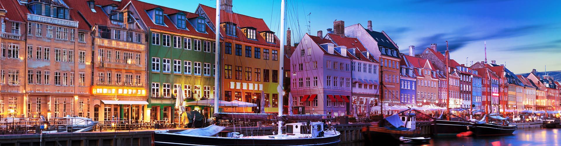 Panoramic view of Copenhagen, Denmark, at dusk, featuring colorful historic waterfront buildings, boats, and illuminated harbor reflections.