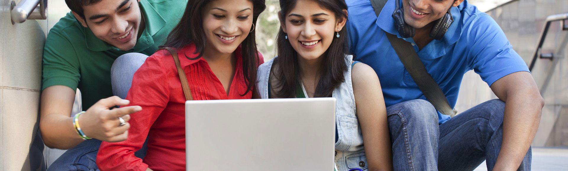 Group of young adults smiling and collaborating around a laptop outdoors.
