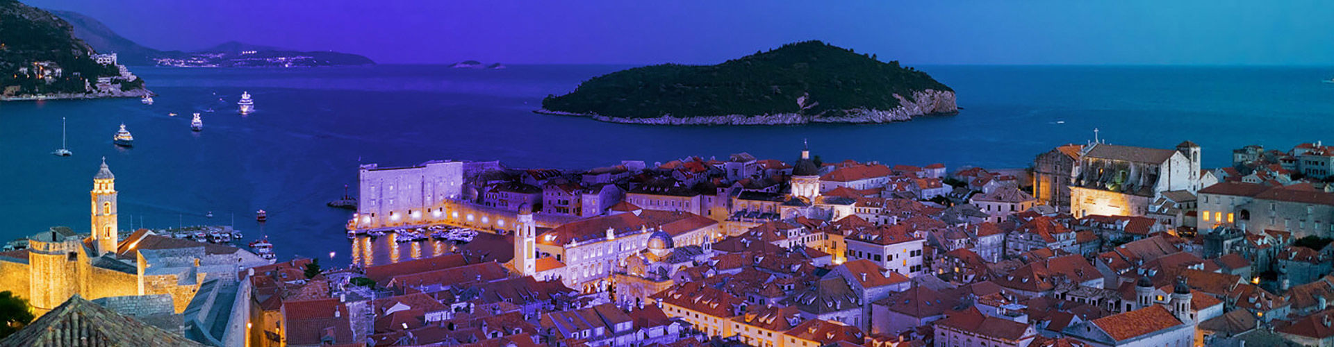 Aerial view of Dubrovnik's historic old town in Croatia at dusk, with the walled city's red-tiled rooftops illuminated, the old harbor below, and the forested Lokrum island in the background under a deep blue-violet sky.