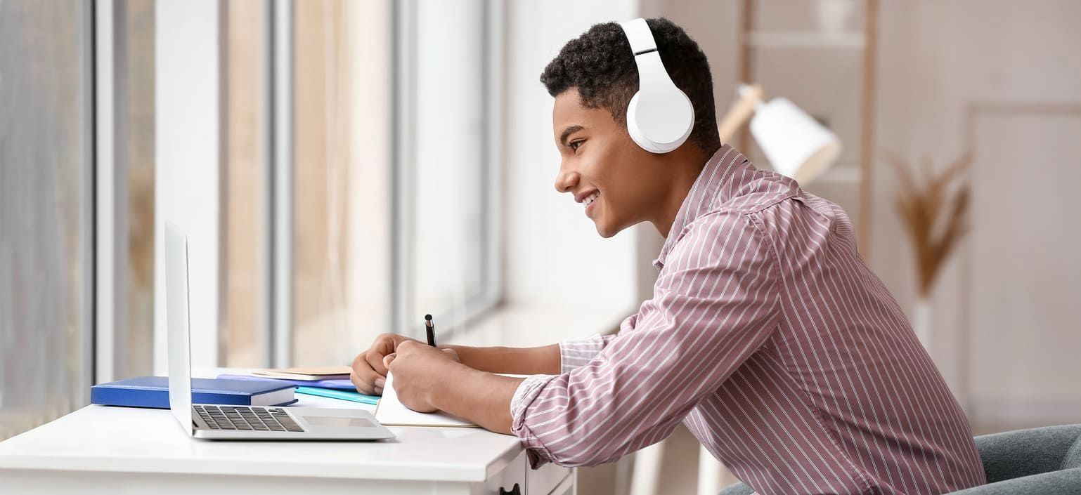 a man sat at desk with a laptop open, whilst wearing headphones and writing in a book