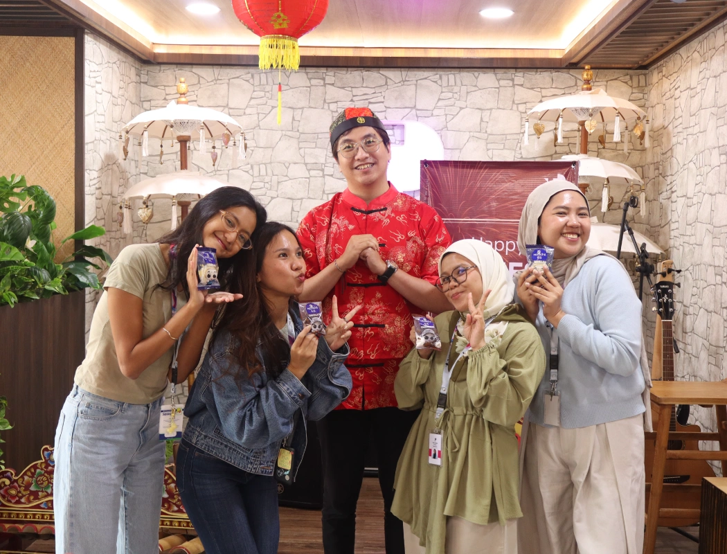 A diverse group of five colleagues poses joyfully during a festive multicultural celebration, blending Chinese and Indonesian cultural elements.
