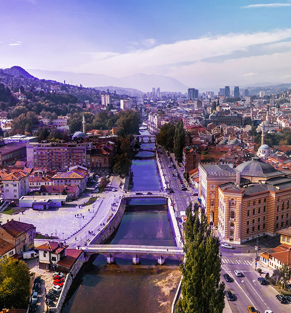 Panoramic view of Sarajevo, Bosnia and Herzegovina, with historic buildings and mosques in the foreground, surrounded by wooded hills and mountains in the distance.