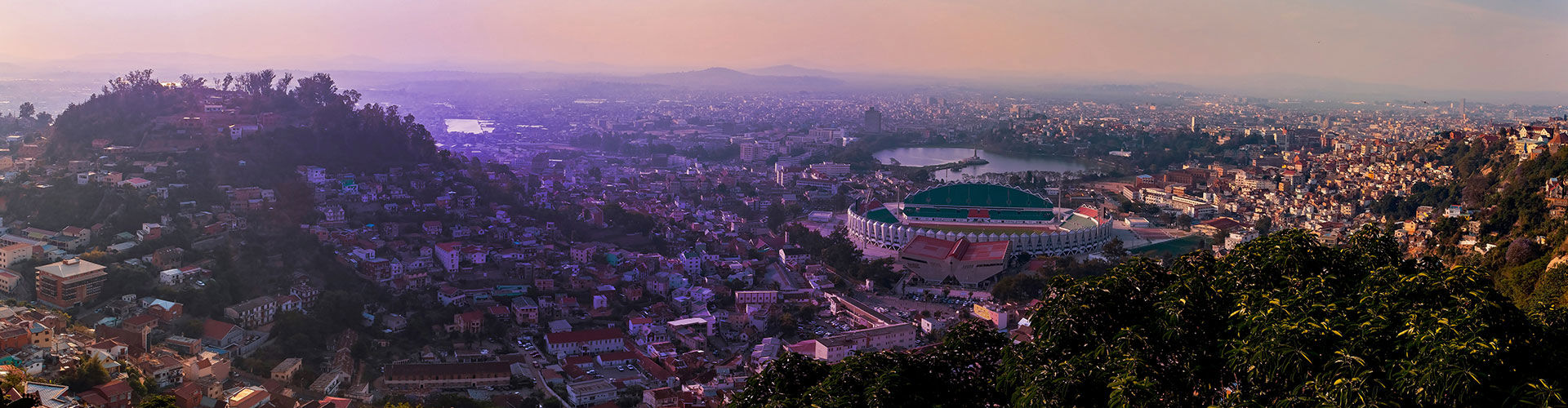 Panoramic view of Antananarivo, Madagascar, at sunset, featuring the city’s dense urban landscape, hills, a large stadium, and mountains in the distance.