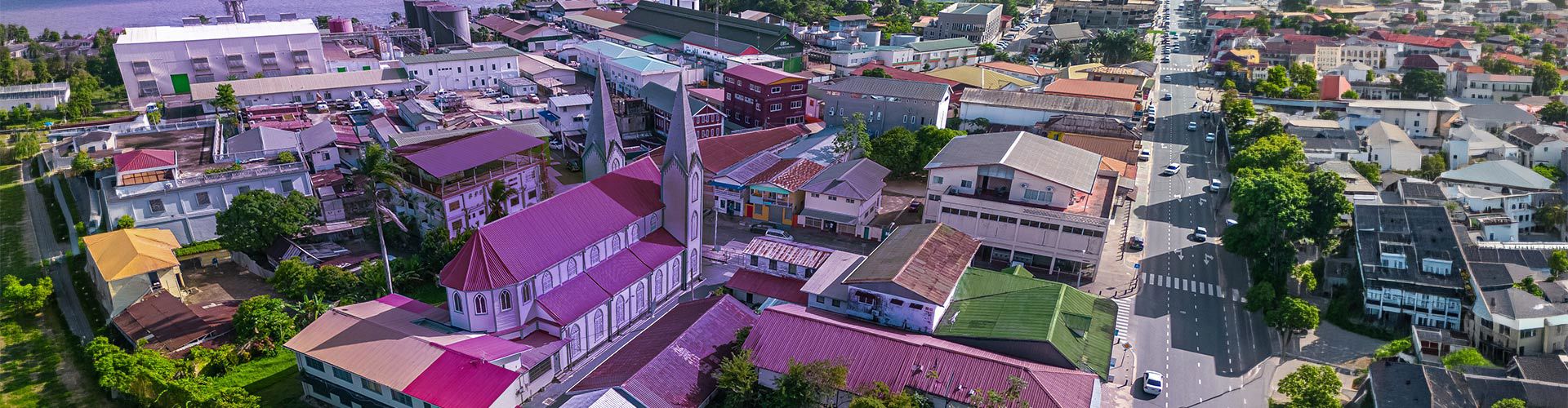 Aerial view of Paramaribo, Suriname, featuring a church with a tall steeple surrounded by colorful rooftops, residential buildings, tree-lined streets, and a waterfront area.