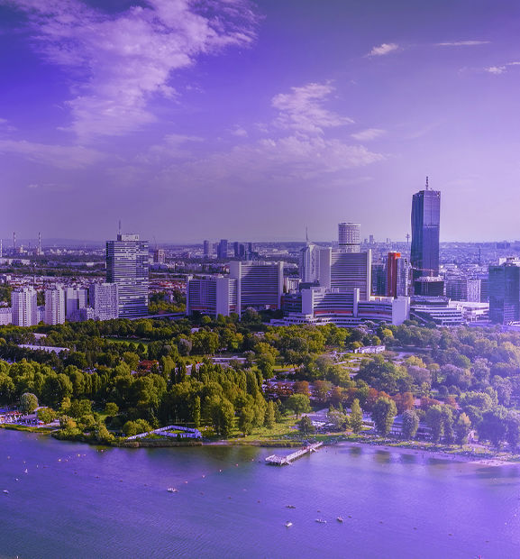 Panoramic aerial view of Vienna, Austria, featuring the modern city skyline, the Danube River, bridges, and lush green parks.