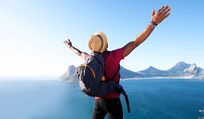 Traveler with arms raised enjoying panoramic view of blue ocean and rugged mountains from a high cliff, wearing a straw hat and backpack under clear sunny sky.