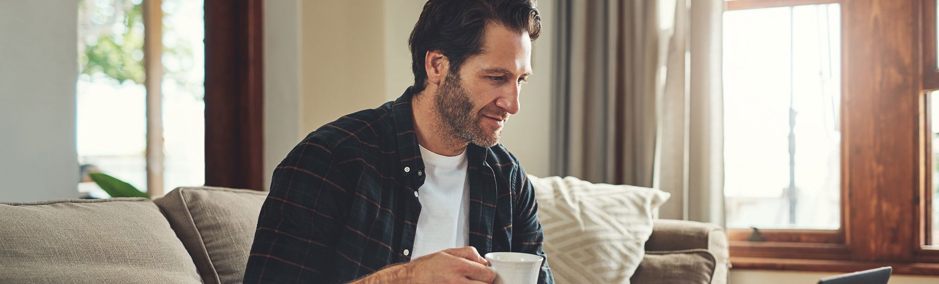 Man sitting on a sofa at home, holding a coffee mug while looking at a laptop.