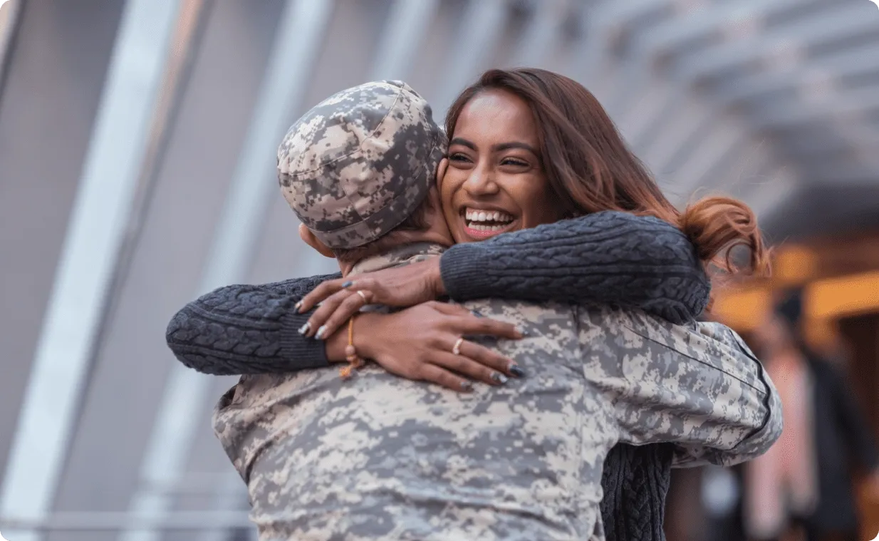 A person in military uniform embraces a smiling woman, both sharing a joyful hug in an outdoor urban setting with blurred architectural elements in the background.