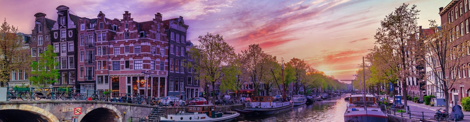Canal view in Amsterdam, Netherlands, at sunset, featuring historic narrow houses, bicycles, boats, and tree-lined streets.