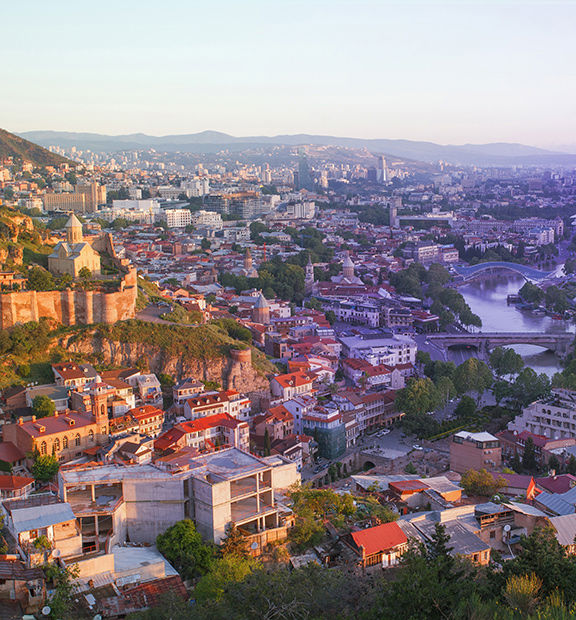 Panoramic view of Tbilisi, Georgia, featuring the historic old town, Narikala Fortress, the Kura River, modern architecture, and mountains in the background.