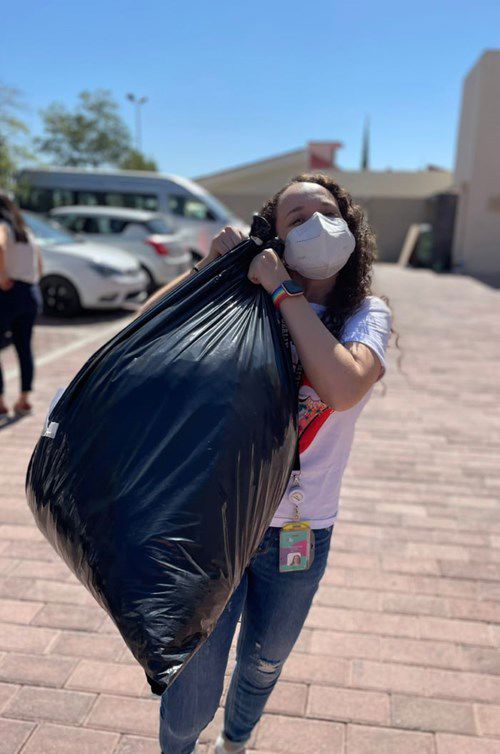 a person wearing a mask carries a large black garbage bag over their shoulder, walking on a paved area in a sunny outdoor setting with parked cars in the background.