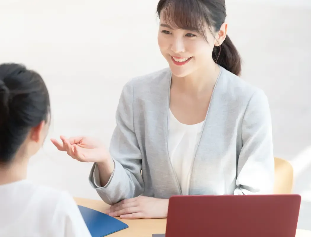 A smiling professional woman gestures while conversing with a colleague at a desk.