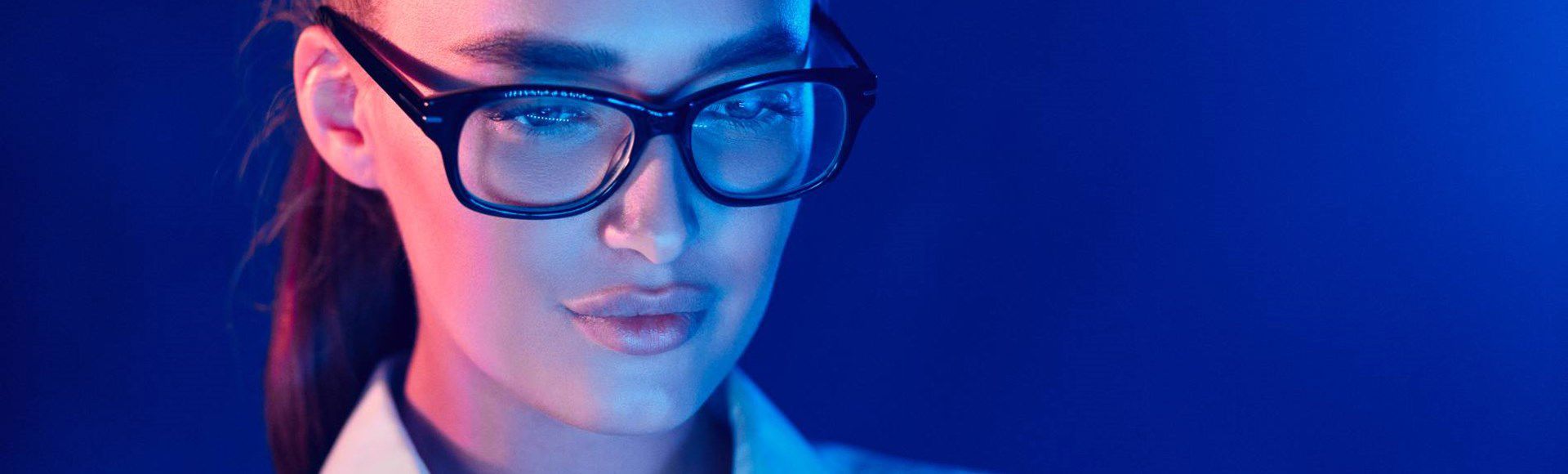 A person wearing glasses and a white shirt, illuminated by blue and pink lighting, appears focused and engaged, set against a solid blue background.