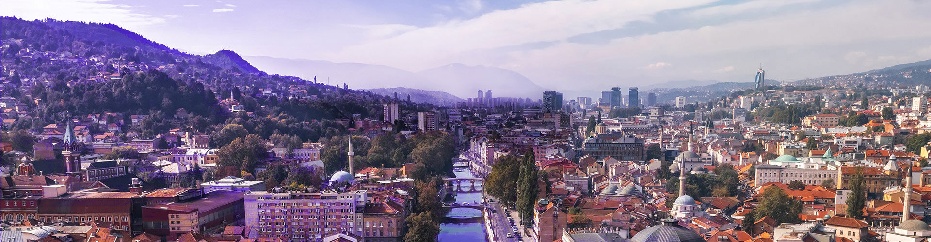 Panoramic view of Sarajevo, Bosnia and Herzegovina, with historic buildings and mosques in the foreground, surrounded by wooded hills and mountains in the distance.