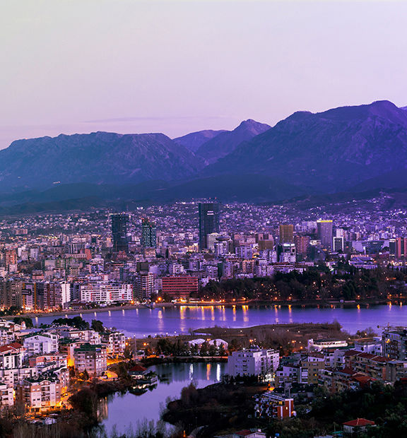 Panoramic view of Tirana, Albania, at dusk, featuring a dense city skyline, modern buildings, a waterfront park, and mountains in the background.