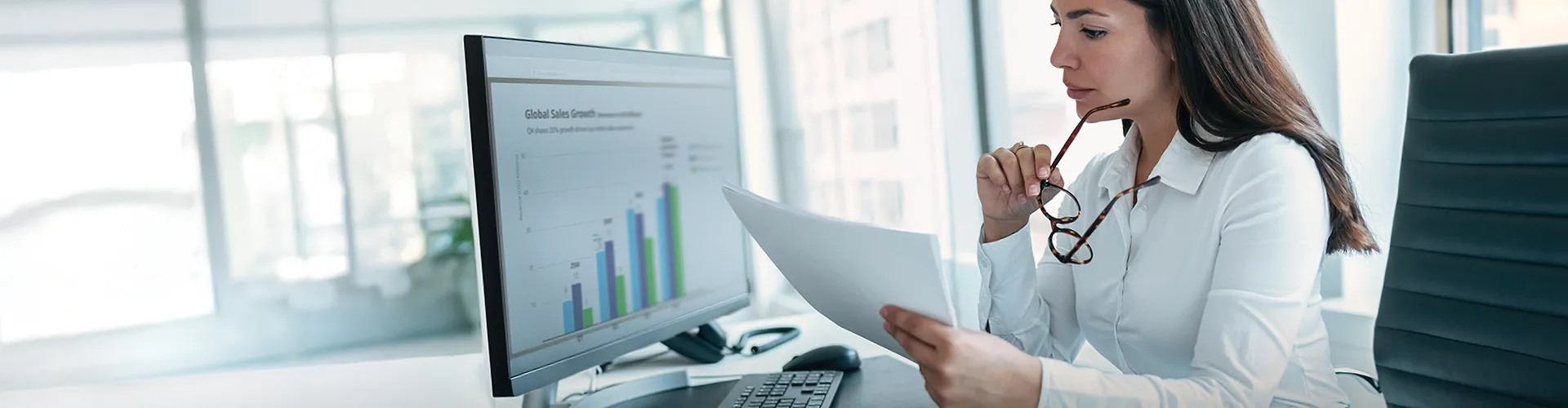 Businesswoman reviewing a report while analyzing sales data on a computer screen.