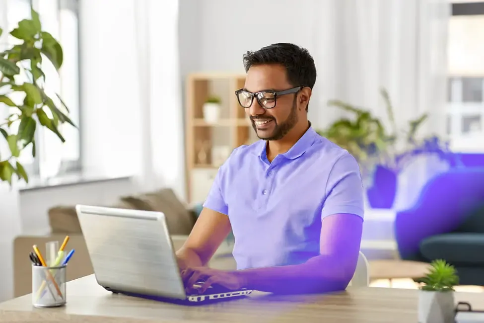 Man wearing glasses and a yellow shirt working on his laptop in a bright, plant-filled home.