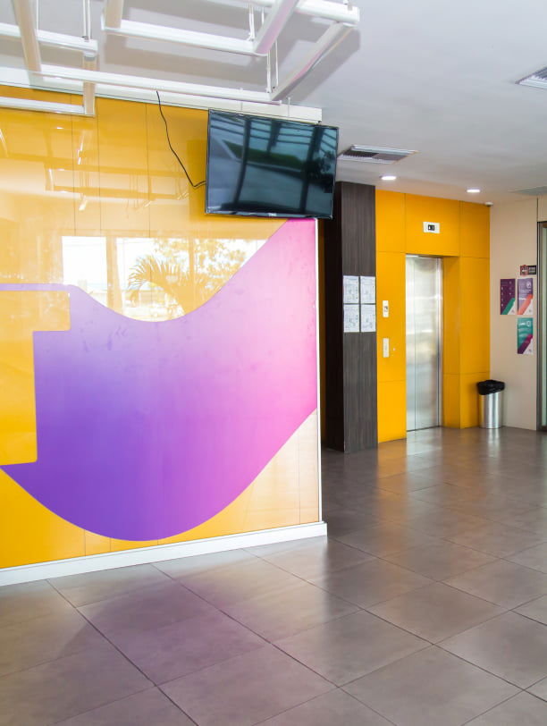 Área de recepción con paredes amarillas, un mural abstracto morado y pantallas montadas en la pared cerca de una puerta.