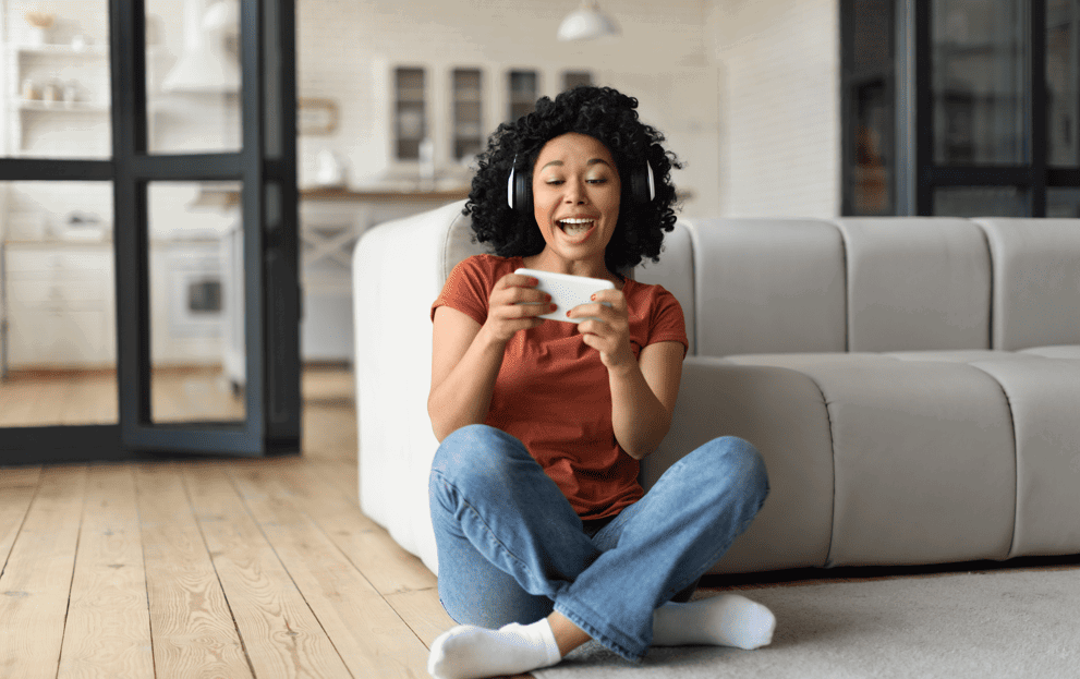 a person wearing headphones plays a game on a smartphone, sitting on a wooden floor beside a gray sofa in a bright, modern living room with a kitchen visible in the background.