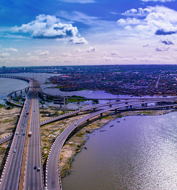Aerial view of Lagos, Nigeria, showing expansive highways and bridges crossing waterways, with the city stretching toward the horizon under a partly cloudy sky.