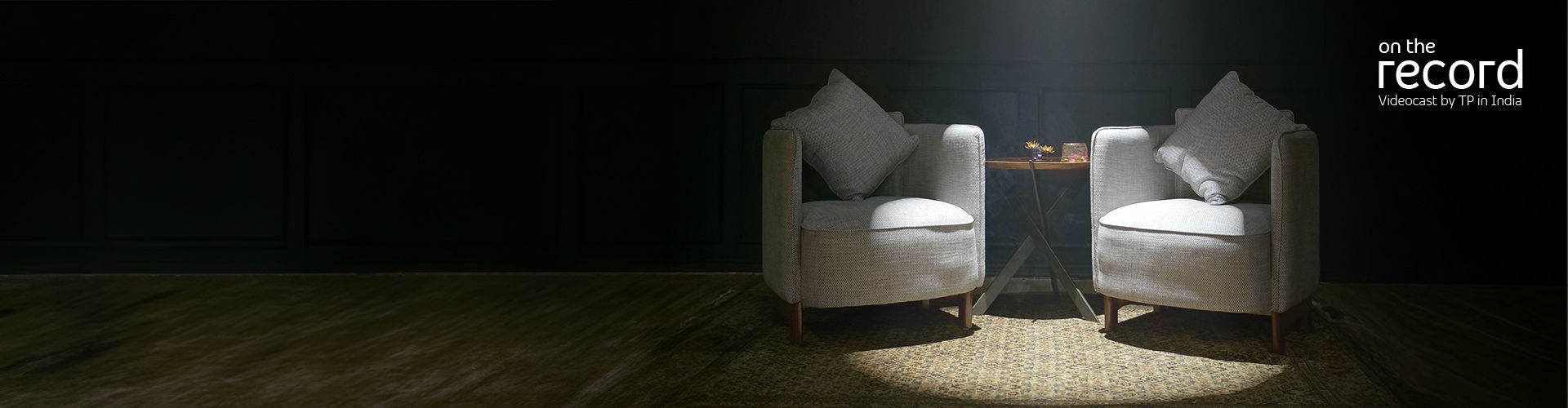Studio with two armchairs and a small table on a rug under spotlight; dark paneled wall background.