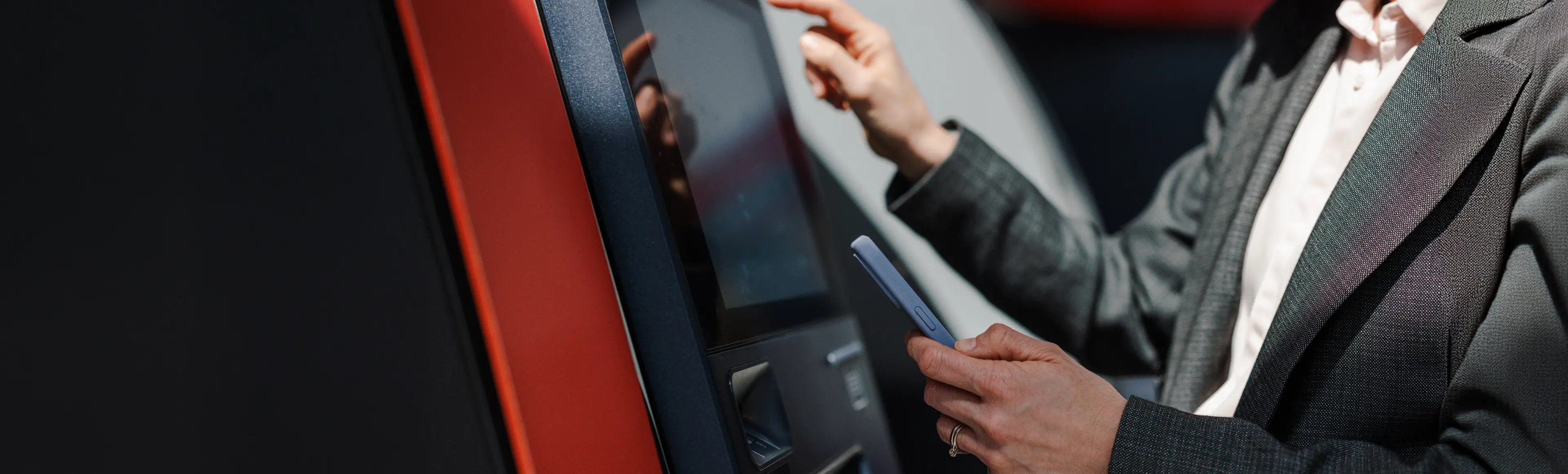 Person using a self-service banking kiosk while holding a smartphone, highlighting TP's digital banking solutions.