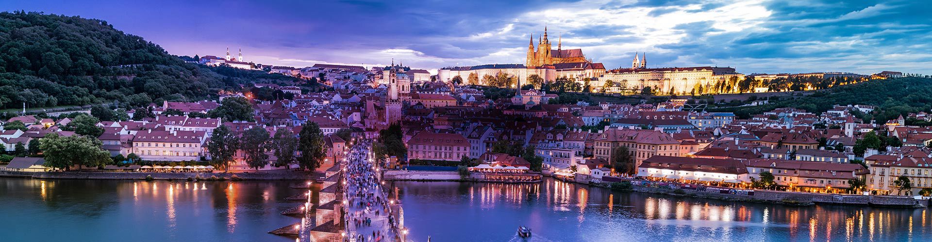 Panoramic view of Prague, Czech Republic, at dusk, featuring Prague Castle, the historic city skyline, Charles Bridge, and the illuminated Vltava River.