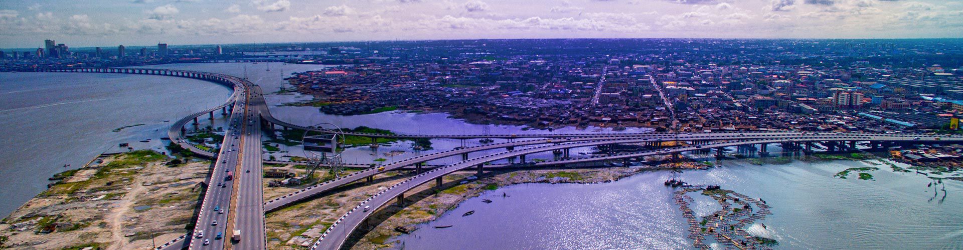 Aerial view of Lagos, Nigeria, showing expansive highways and bridges crossing waterways, with the city stretching toward the horizon under a partly cloudy sky.