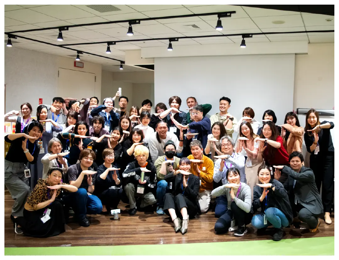 A large diverse group poses playfully with matching hand gestures indoors.