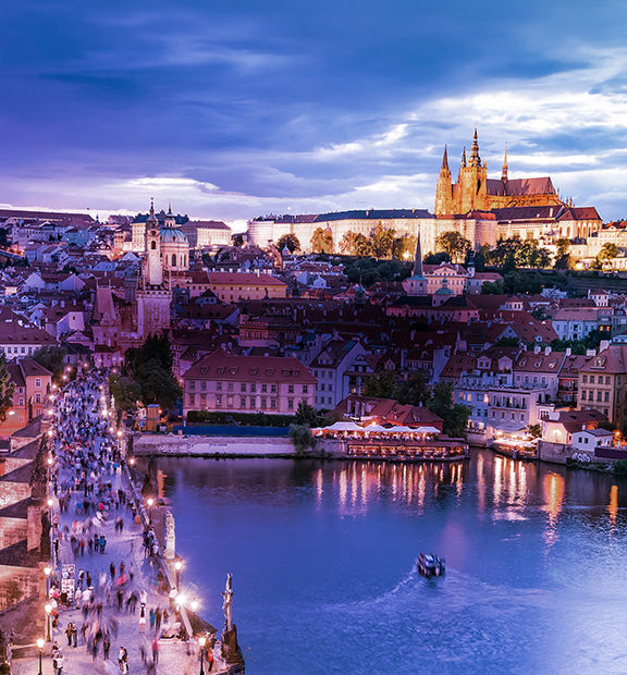 Panoramic view of Prague, Czech Republic, at dusk, featuring Prague Castle, the historic city skyline, Charles Bridge, and the illuminated Vltava River.