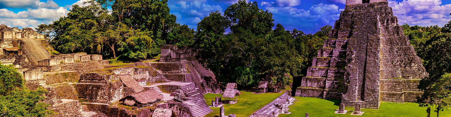 Panoramic view of the ancient Maya ruins at Tikal, Guatemala, surrounded by lush tropical forest, with towering stone temples, plazas, and historic structures under a partly cloudy sky.