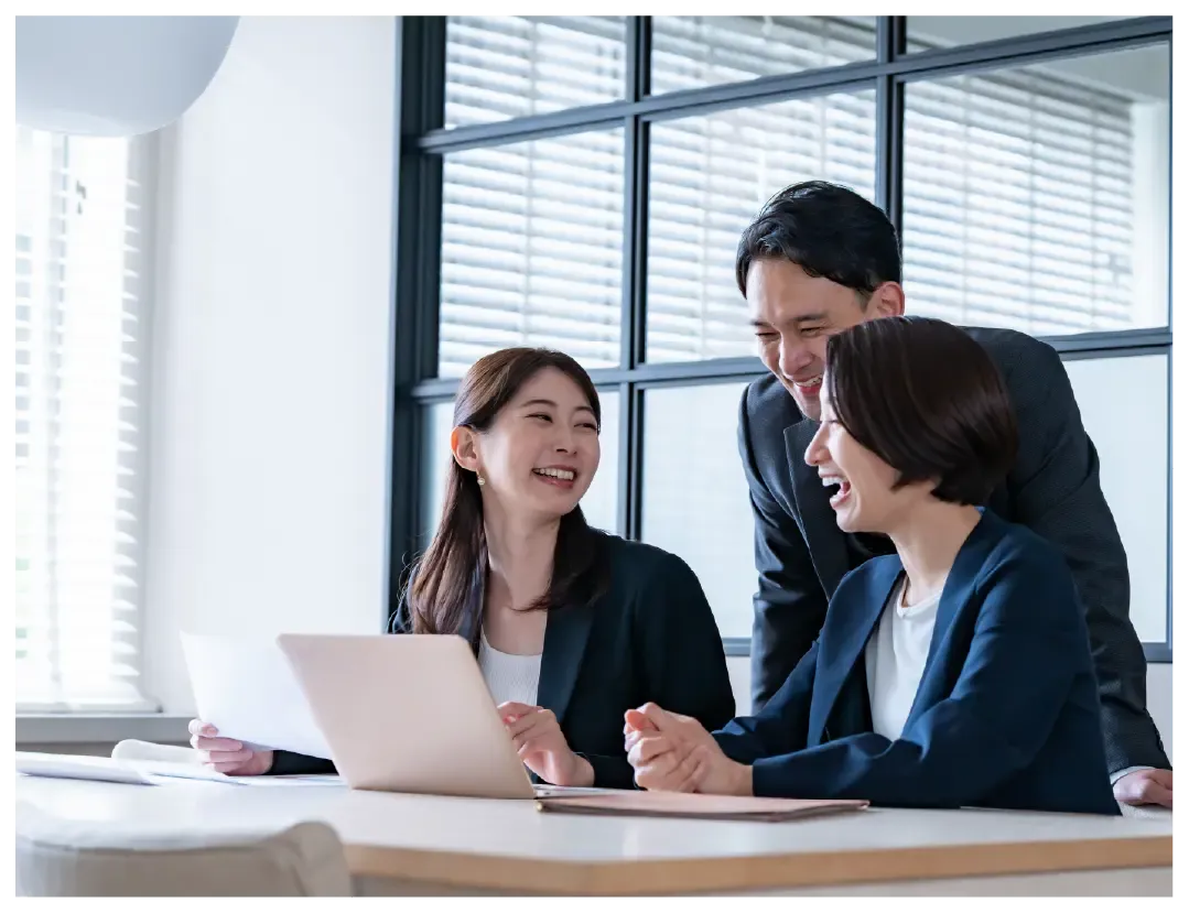 Three colleagues laugh together while collaborating at a bright office desk.