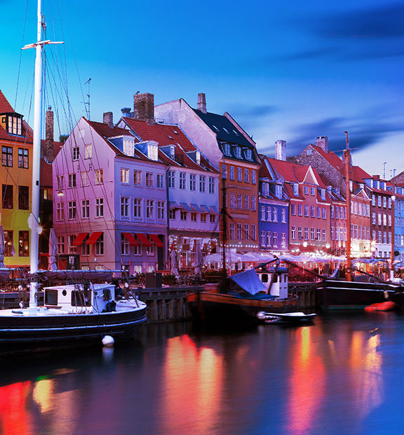 Panoramic view of Copenhagen, Denmark, at dusk, featuring colorful historic waterfront buildings, boats, and illuminated harbor reflections.